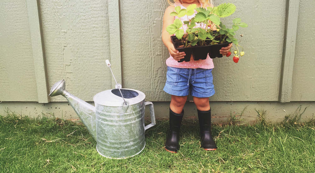 A girl is holding gardening plants with a metal watering can for her mud kitchen.