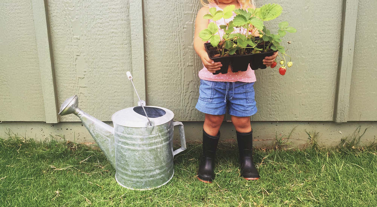 A girl is holding gardening plants with a metal watering can for her mud kitchen.