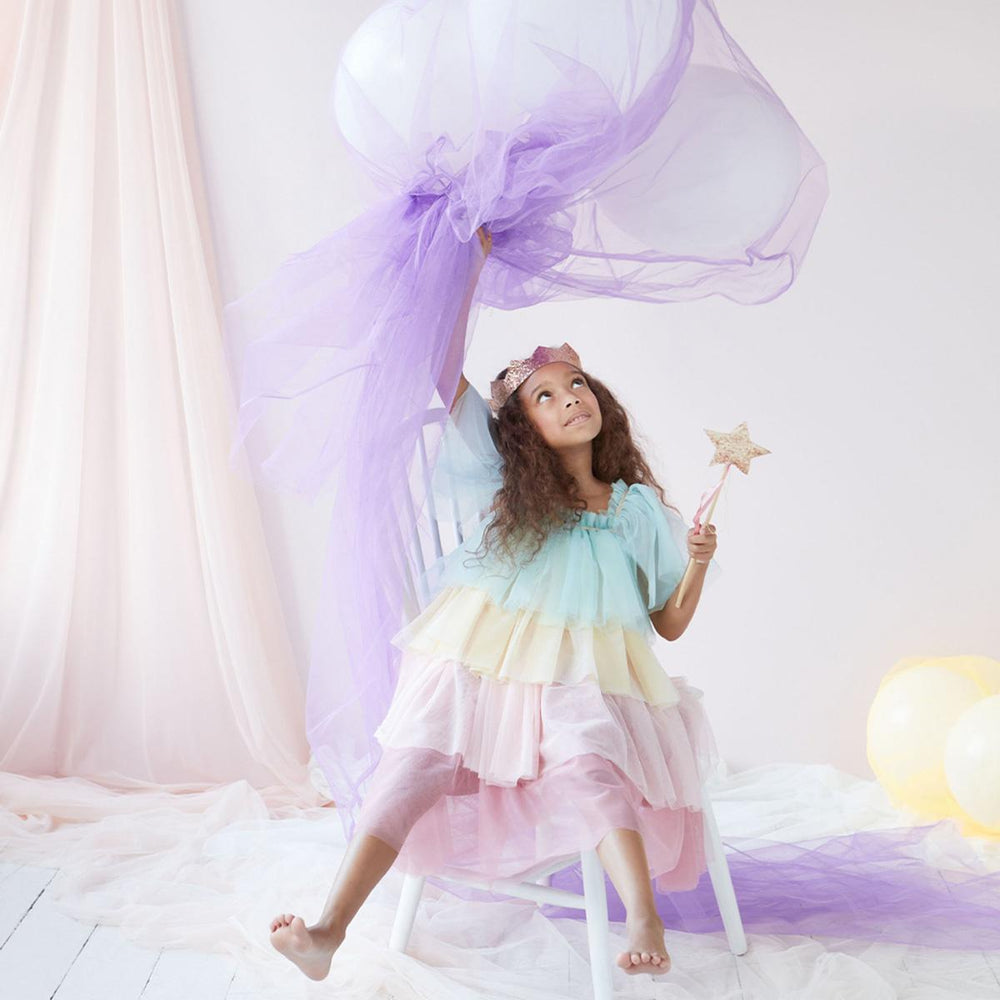 Young child wearing Meri Meri's pastel rainbow tulle dress and gold crown, sitting on a stool and waving sheer purple fabric and holding a gold star wand in the other hand.