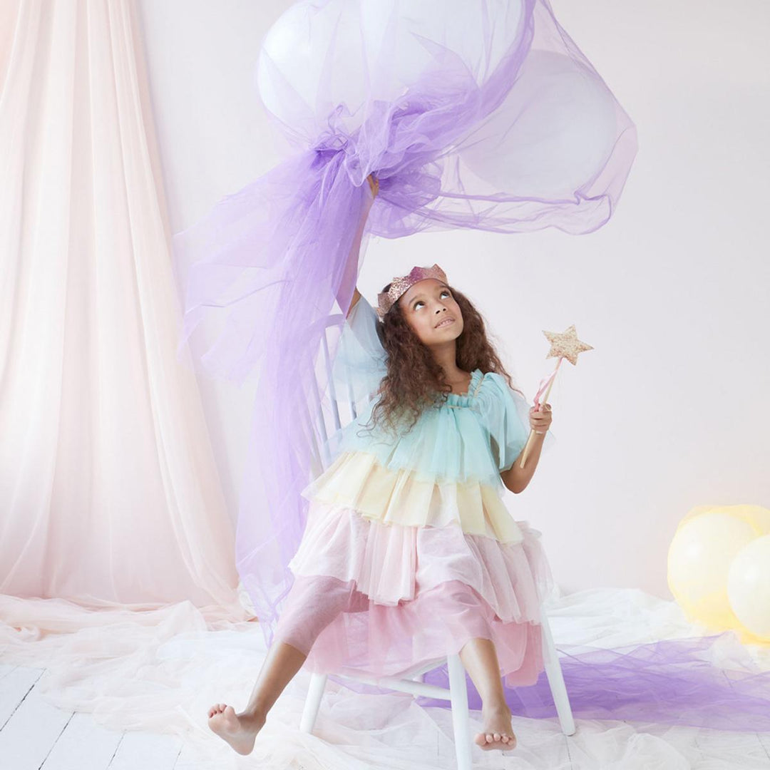 Young child wearing Meri Meri's pastel rainbow tulle dress and gold crown, sitting on a stool and waving sheer purple fabric and holding a gold star wand in the other hand.