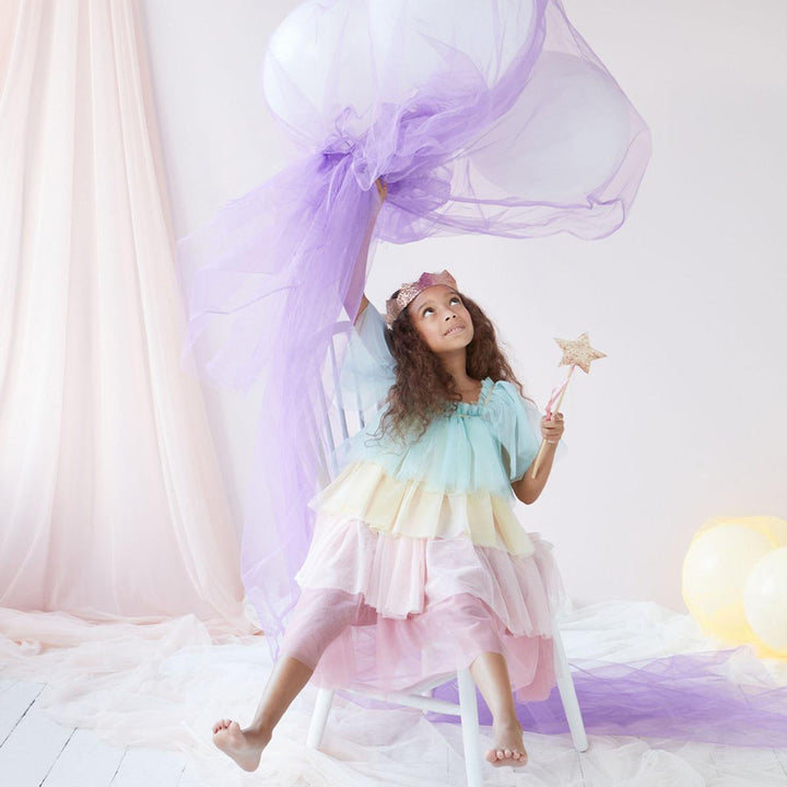 Young child wearing Meri Meri's pastel rainbow tulle dress and gold crown, sitting on a stool and waving sheer purple fabric and holding a gold star wand in the other hand.