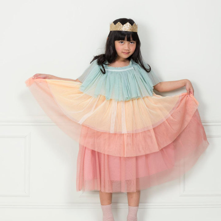 Child wearing the children’s pastel Rainbow Ruffle Princess Costume Set featuring a rainbow tulle dress with large flutter sleeves and gold glitter crown from Meri Meri standing against a white wall and holding the skirt out in their hands.
