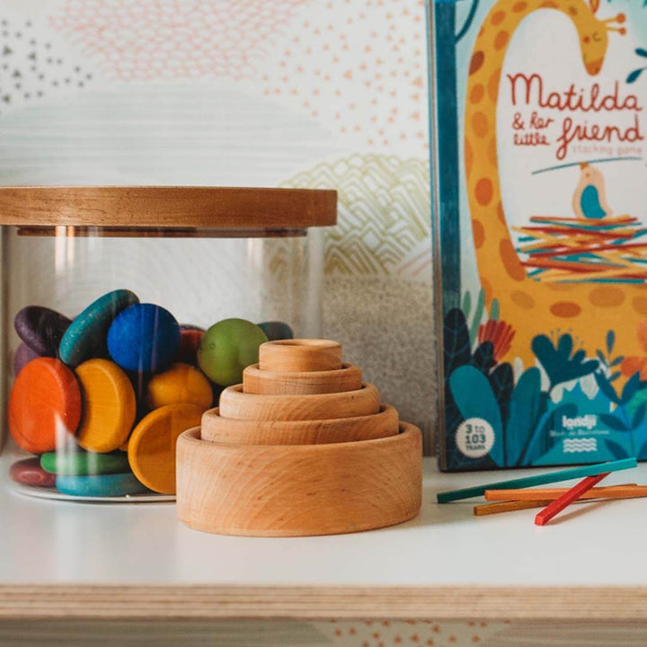 Wooden stacking bowls and colorful wooden blocks on a shelf with a book titled 'Matilda & her little friend' in the background.