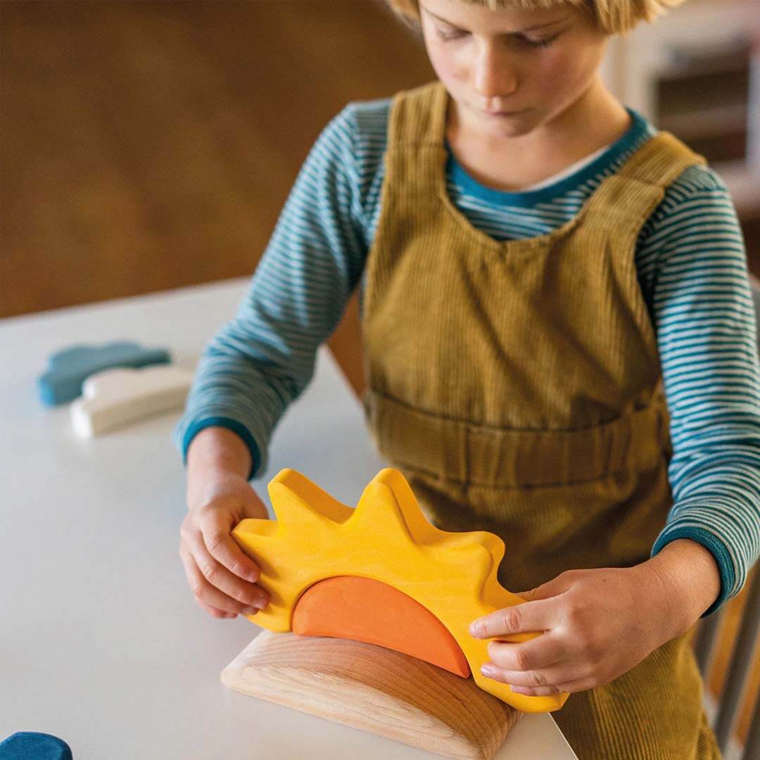 Child wearing a brown apron holding a yellow and orange sun-shaped wooden toy.