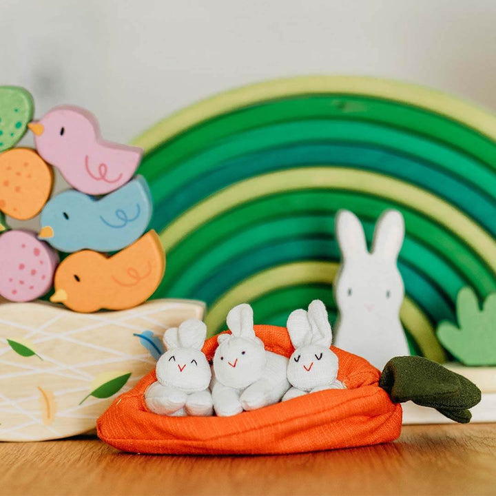 Toy rabbits on a carrot with a wooden rainbow and birds in the background.