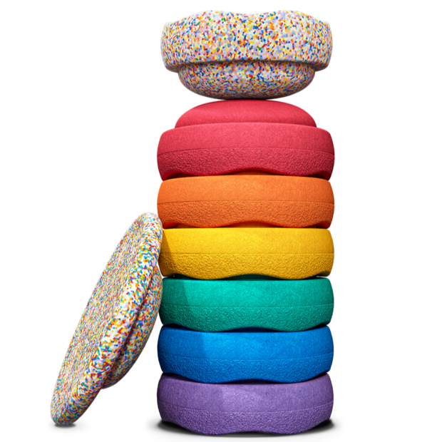 Stack of seven colorful Stapelstein® stepping stones in rainbow colors with a Super Confetti element on top and one Balance Board resting on the side—complete set for active, open-ended play.