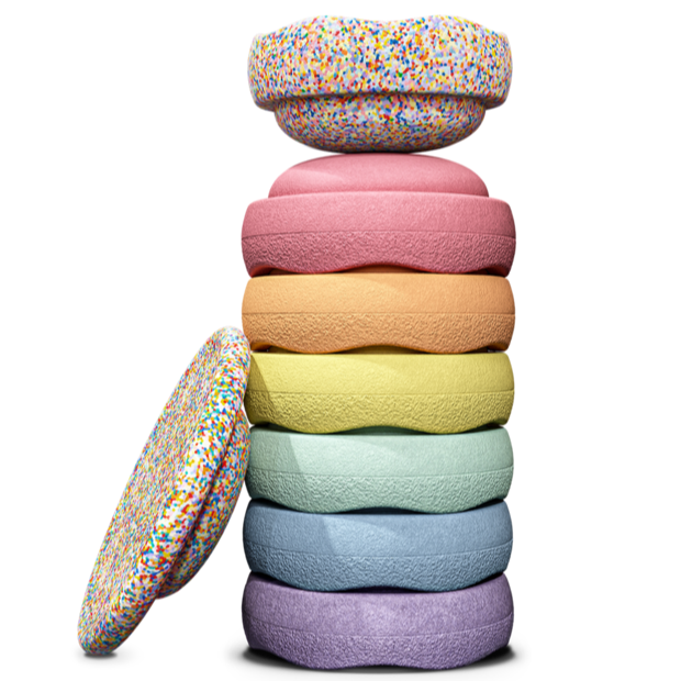 Stack of seven colorful Stapelstein® stepping stones in pastel rainbow colors with a Super Confetti element on top and one Balance Board resting on the side—complete set for active, open-ended play.