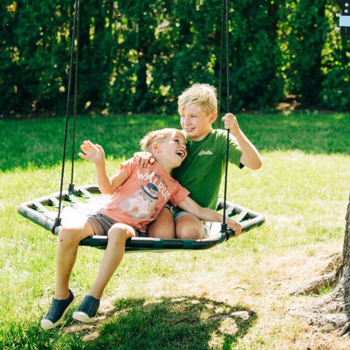 Two children swing on a Bijou Build Kubix hexagon swing in the backyard.