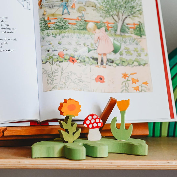 Wooden toy with flowers and a mushroom on a table in front of an open book with illustrations.