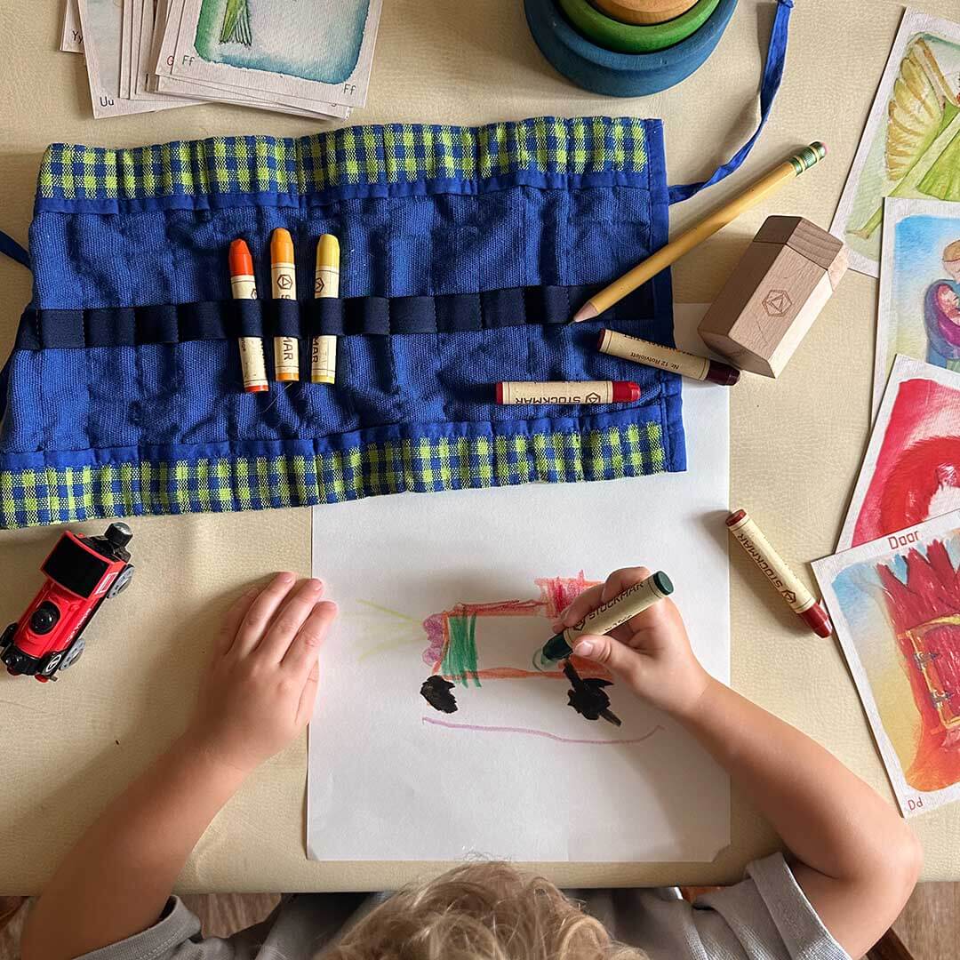Child drawing with markers on a piece of paper at a table with art supplies.