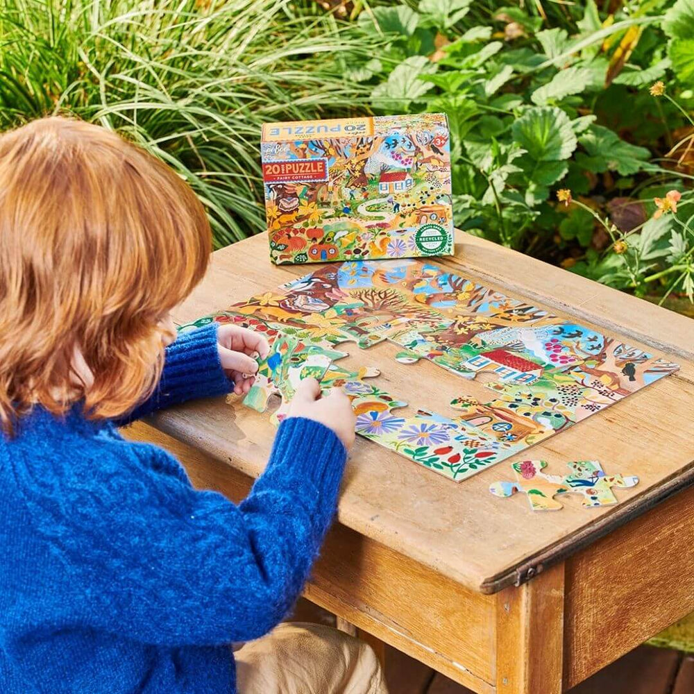 Child in a blue sweater seated at a wooden table in a garden assembling the eeBoo Fairy Cottage 20-Piece Puzzle featuring a whimsical woodland scene with fairies, animals, and a cozy cottage, for kids aged 3+.