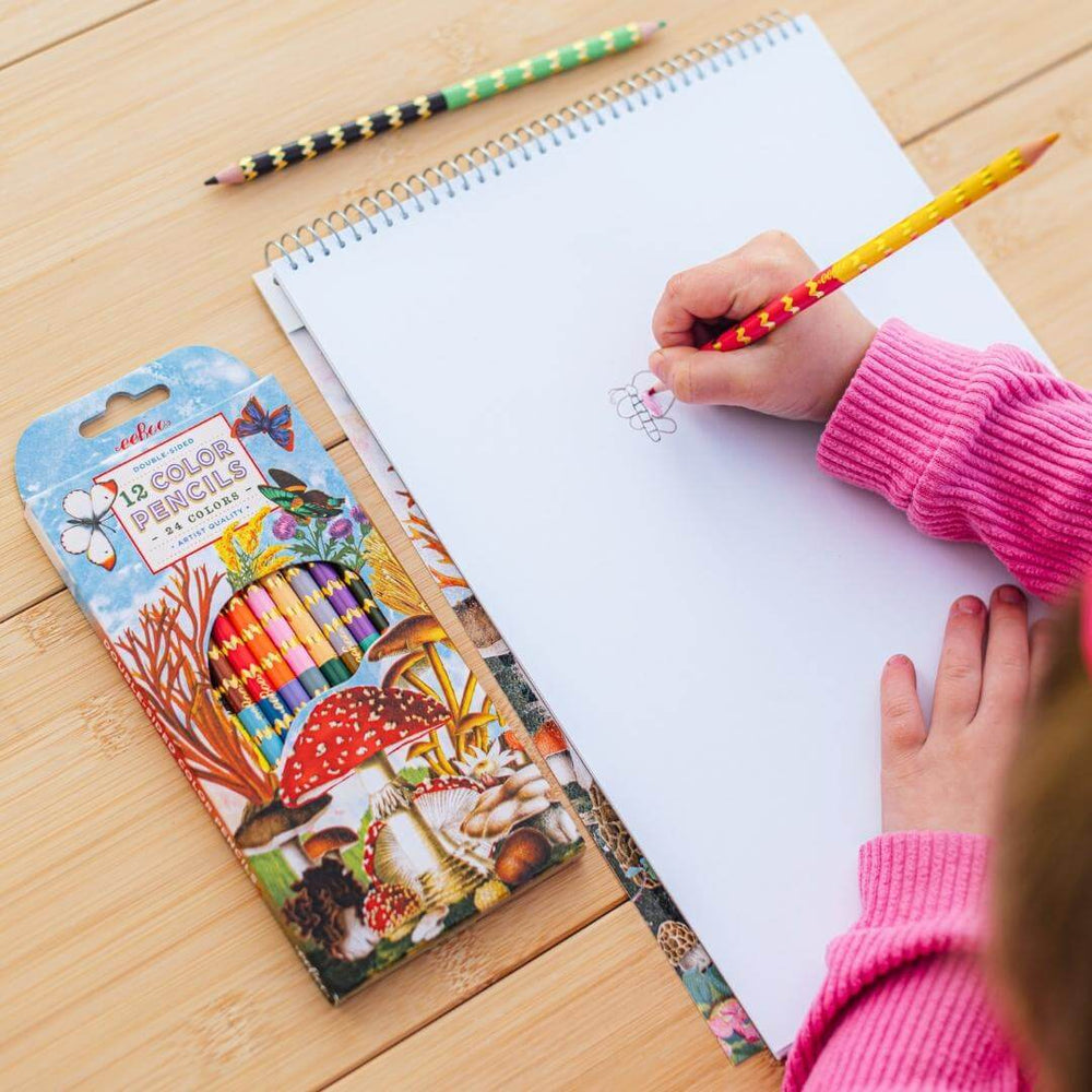 Child drawing in eeBoo Mushroom Sketchbook featuring vibrant mushroom and butterfly collage artwork by Fumiha Tanaka. Spiral-bound pad with 60 matte pages, on a wooden table.
