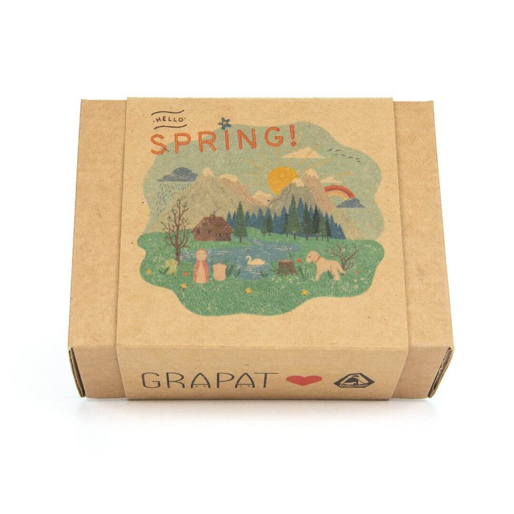 A beautifully illustrated cardboard box from the Grapat x Ostheimer 'Hello Spring' set. The packaging features a whimsical nature scene with rolling green hills, a bright sun, trees, and woodland animals, along with the words 'Hello Spring!' in a playful font. The Grapat logo and a small heart icon are printed on the side, highlighting the brand’s commitment to natural, imaginative play.