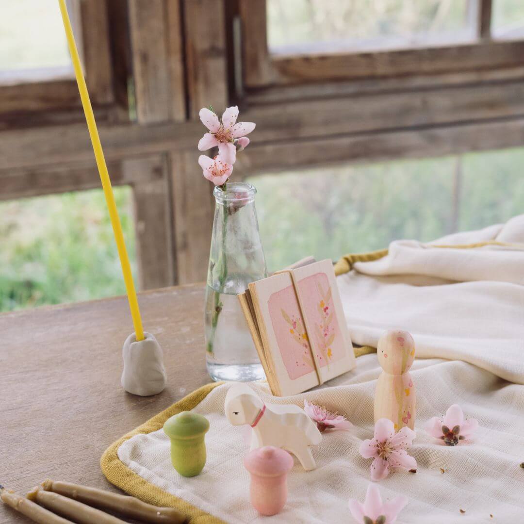 A serene spring-inspired table setting featuring the Grapat x Ostheimer 'Hello Spring' set. The display includes a wooden Nin figure, a miniature Ostheimer lamb, a flower press adorned with blossoms, and small Mandala tulip pieces in pink and green. A glass vase with delicate pink flowers, a lit beeswax candle, and natural linen fabric create a warm and inviting seasonal atmosphere by the window.