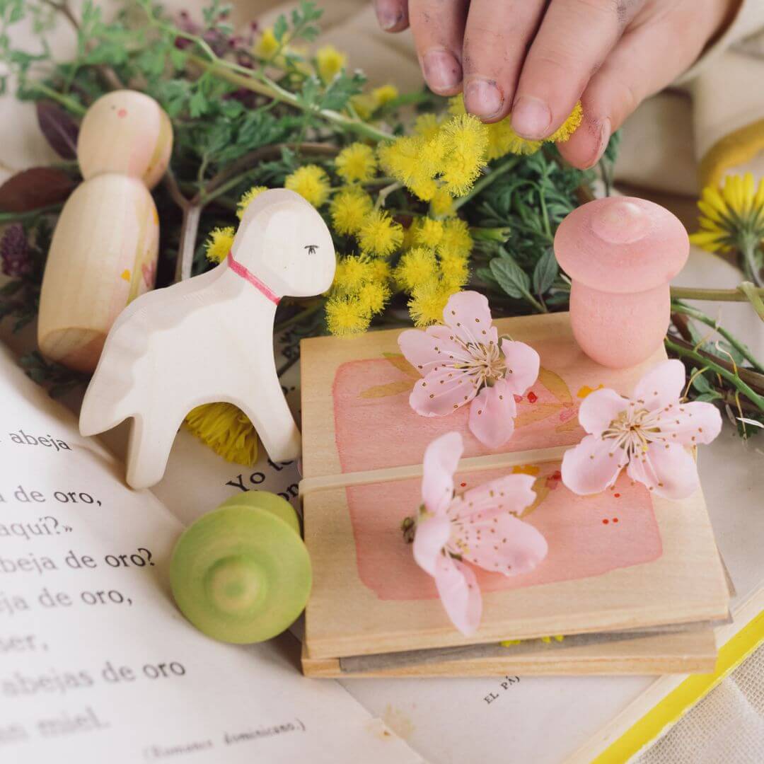 A whimsical spring-themed play setup featuring the Grapat x Ostheimer 'Hello Spring' set. The scene includes a wooden Ostheimer lamb with a pink ribbon collar, a flower press adorned with real blossoms, a wooden Nin figure, and small Mandala tulip pieces in pink and green. Yellow spring flowers and hands arranging natural elements add to the enchanting and nature-inspired atmosphere.