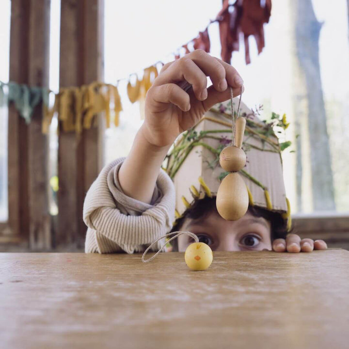 A young child, partially hidden behind a table, curiously reaches up to hang a handcrafted wooden bunny ornament from Grapat. Another small yellow chick ornament rests on the table. The child wears a nature-inspired headband, and the background features a cozy, rustic setting with hanging fabric decorations and natural light.