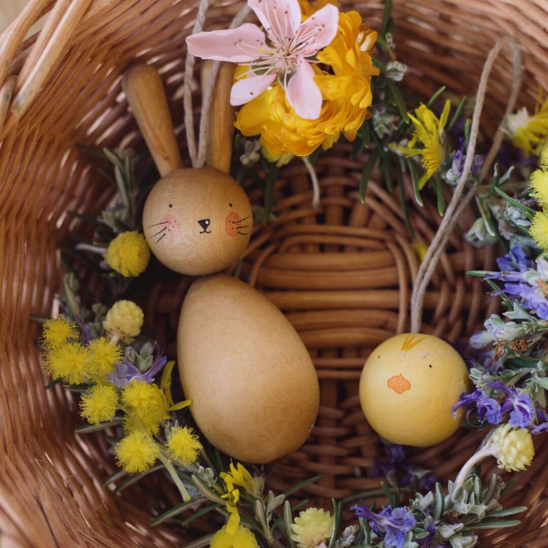 A wicker basket filled with spring flowers and two handcrafted wooden Easter ornaments from Grapat: a bunny with long ears and a small yellow chick. The ornaments are nestled among delicate yellow, purple, and pink flowers, creating a festive and natural Easter display.