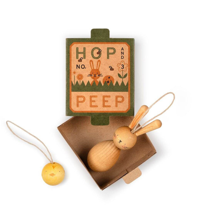 A set of handcrafted wooden Easter ornaments from Grapat, including a small wooden bunny and chick with hanging loops, nestled inside an open box. The packaging features a vintage-inspired design with the words 'HOP' and 'PEEP' along with illustrations of rabbits and chicks. The ornaments are designed for hanging and celebrating the joy of spring and Easter.