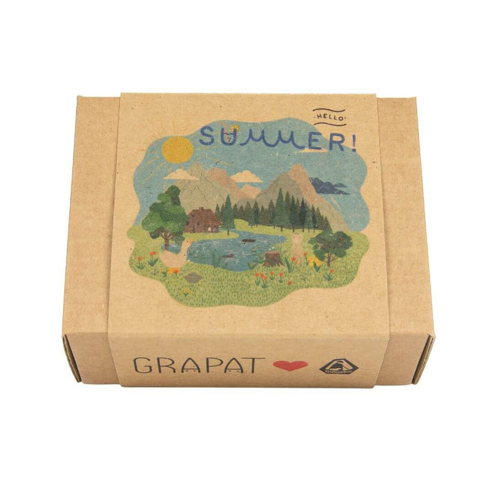 Closed kraft paper box labeled 'Hello Summer!' with whimsical illustrated artwork showing a sunlit landscape of trees, a pond, mountains, and a cozy cabin, marking the seasonal Grapat x Ostheimer collaboration.