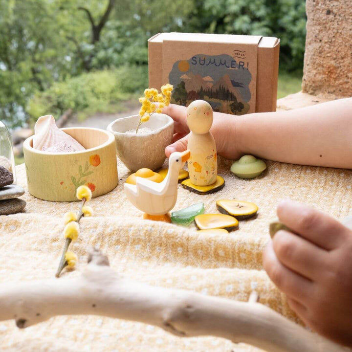 Complete Hello Summer set displayed outdoors with children's hands shown, with wooden Nin, goose, bowl, and mandala pieces arranged on a cloth, capturing the seasonal spirit of the Grapat and Ostheimer collaboration.