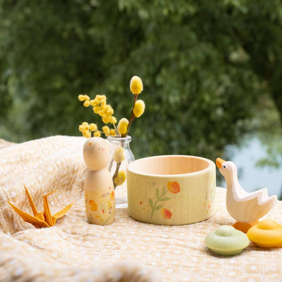 Grapat x Ostheimer Hello Summer collection styled on a soft yellow cloth with fresh flowers. Includes the wooden Nin figure, Ostheimer goose, bowl, and mandala pieces with the illustrated box in the background.