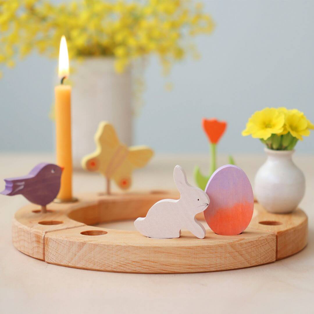 Grimm’s Wooden Easter Egg and Bunny in the Purple Orange Colorway displayed on a wooden celebration ring, accompanied by a yellow candle, a small vase with flowers, a wooden bird, a wooden butterfly, a wooden red tulip, and festive decorations.
