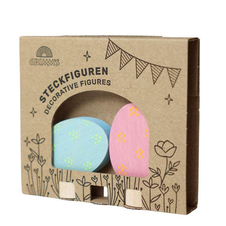 Grimm’s Wooden Easter Eggs in the Floral Colorway, showing thepink with neon orange dotted egg and the pale blue with neon yellow colors of the two eggs in the beautifully illustarted cardboard packaging that they arrive in.