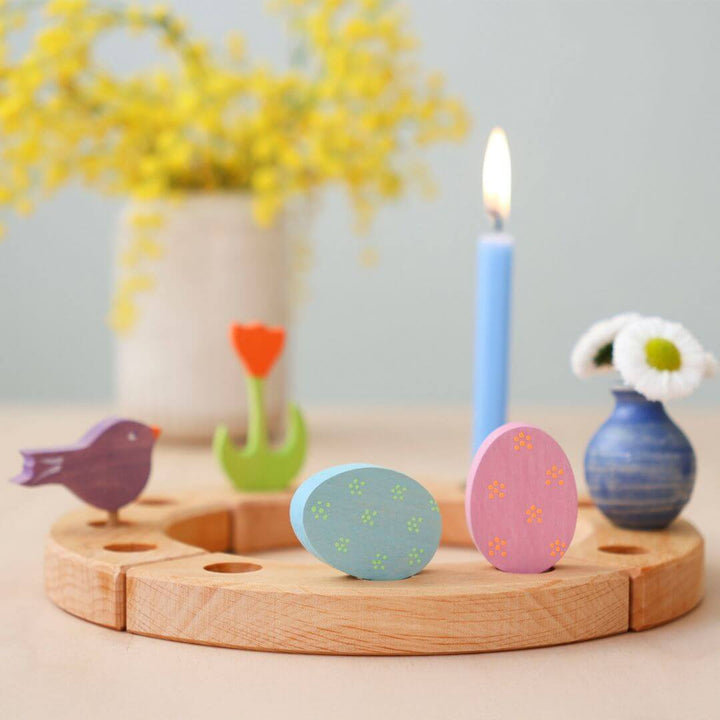 Grimm’s Wooden Easter Eggs in the Floral Colorway displayed on a wooden celebration ring, accompanied by a blue candle, a small vase with flowers, a wooden bird, a wooden red tulip, and festive decorations.