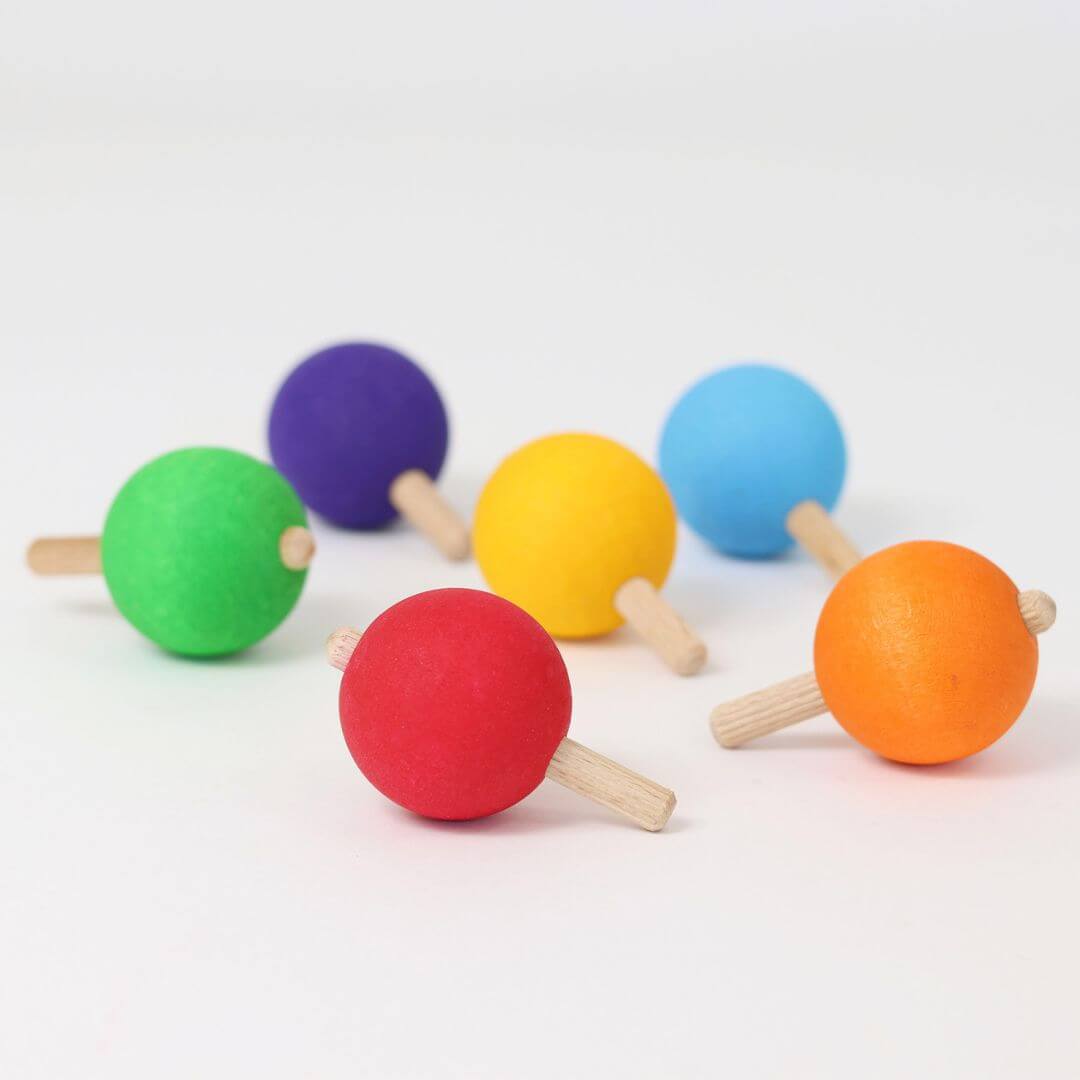 Six colorful wooden spinning tops arranged on a white background, each with a round ball handle, crafted by Grimm’s.