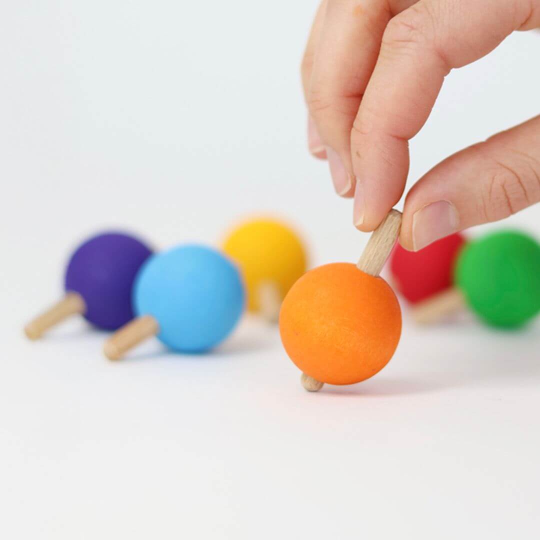 Close-up of a hand spinning an orange wooden rainbow top, with other colorful Grimm’s spinning tops resting in the background.