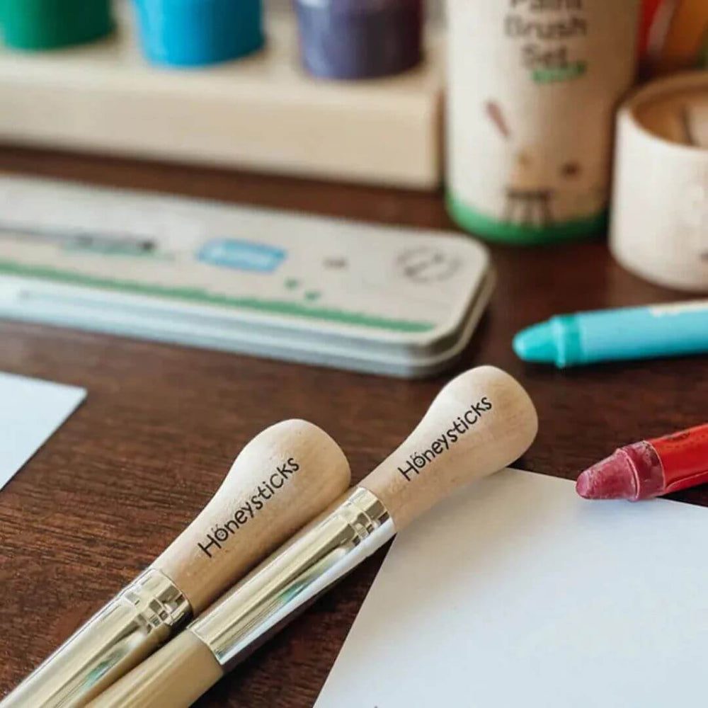 Two 'Honeysticks' paint brushes on a wooden surface with art supplies in the background.