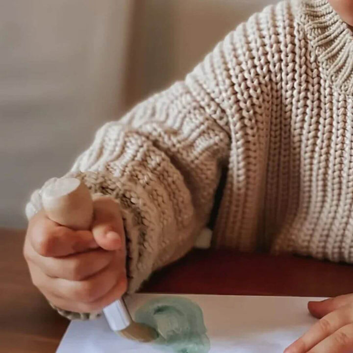 A toddler wearing a beige knit sweater painting with a chunky Honeysticks paintbrush.