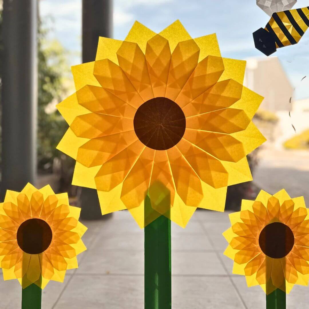 Three folded Japanese silk paper sunflowers on green stems with a blurred outdoor background.