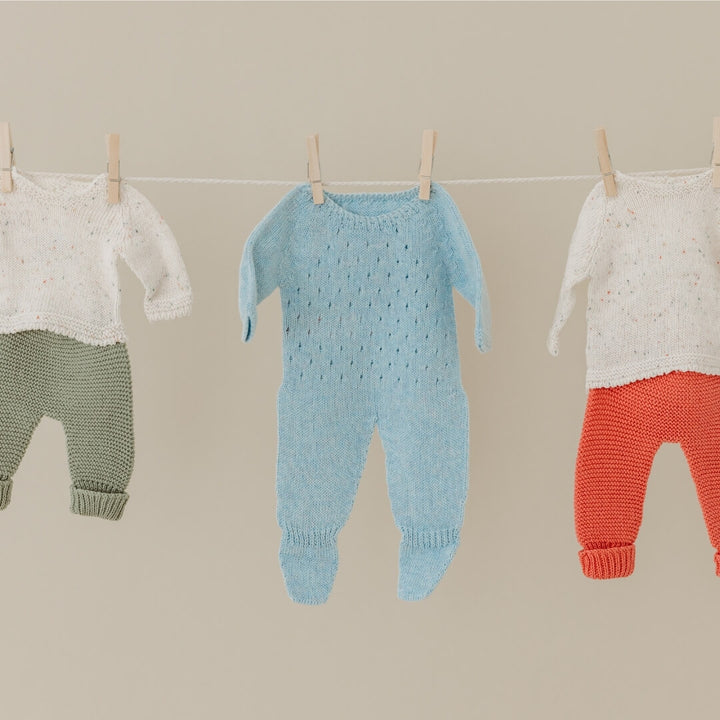 Three knitted baby doll outfits hanging on a line against a beige background including the blue knit pajamas for 15-inch dolls, made from recycled materials with a handmade look—part of Miniland's eco-friendly doll clothing collection.