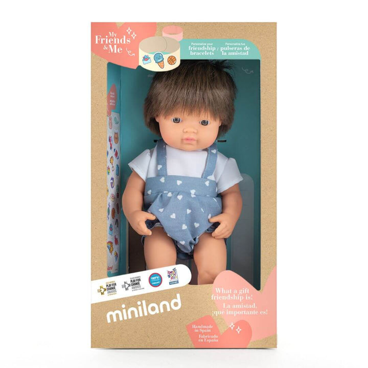 A realistic white boy doll with brown hair, dressed in a light blue romper with white heart patterns, standing up in the box he is packaged in. 