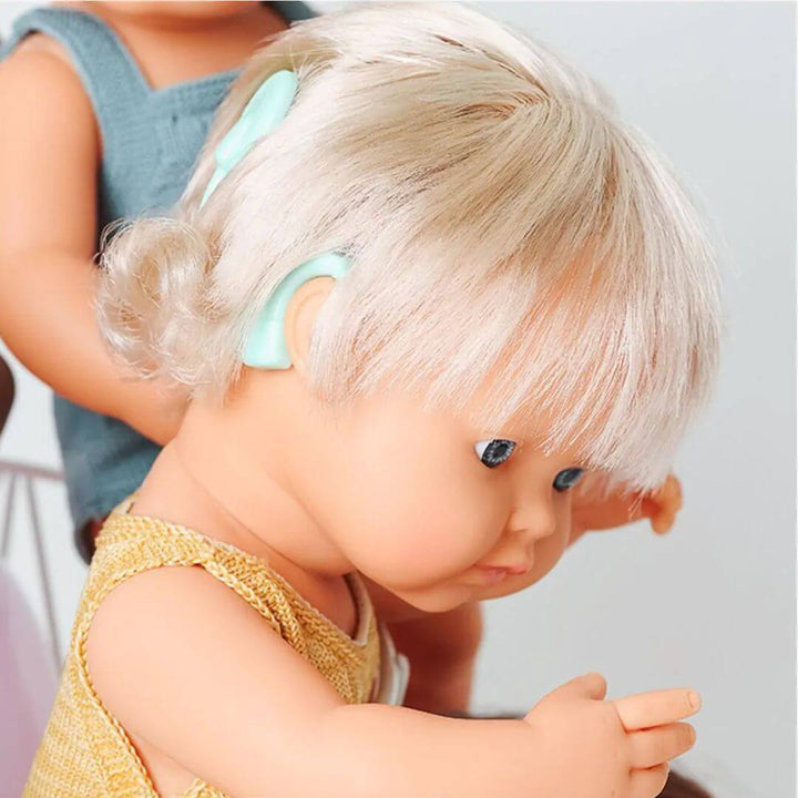 A close up of the cochlear implant on this Miniland Montessori doll with blonde hair wearing a yellow knit top and another doll in the background.