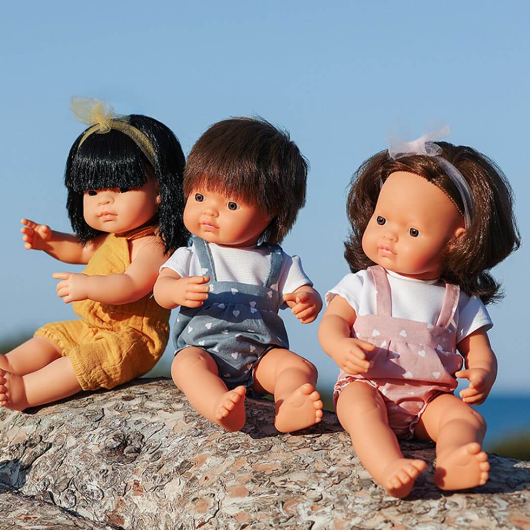 Three Miniland Montessori dolls sitting on a sunlit rock outdoors; the dolls include an Asian girl in a mustard romper, a white boy with brown hair in blue overalls, and a white girl with brown hair in a pink outfit and headband.