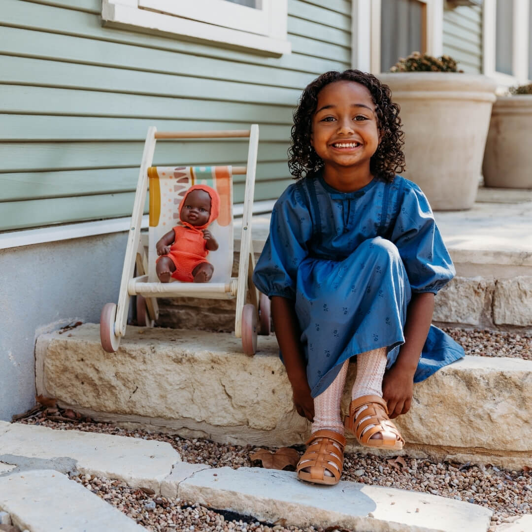 Child in a blue dress sitting on stone steps outside of a house next to a doll in a wooden doll stroller by Miniland, made in Spain from sustainable wood with a colorful fabric seat.