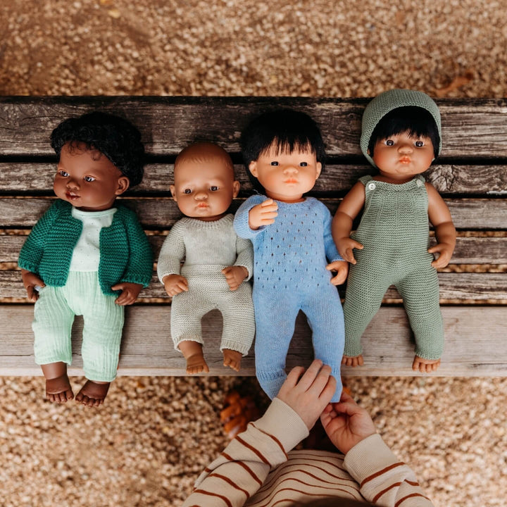 Four Miniland Realistic dolls in knitted outfits on a wooden surface with a hand reaching towards one of them.