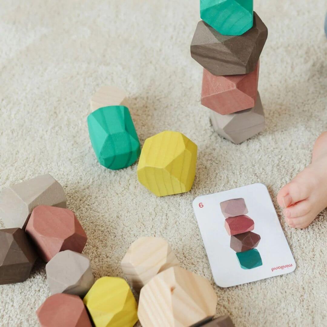 Miniland - Child stacking wooden stones in soft natural tones, exploring balance and tactile play on a cozy rug with a template card on view - Bella Luna Toys