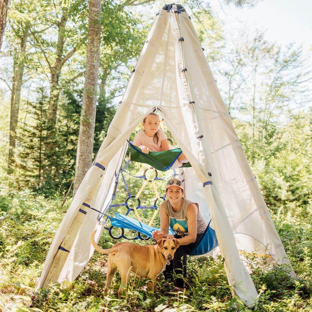 A woman and a child are camping outdoors in a forest with a dog, using a large beige teepee-style shelter with built-in hammocks and a climbing frame. The scene is bright and natural, with trees and grass visible in the background.