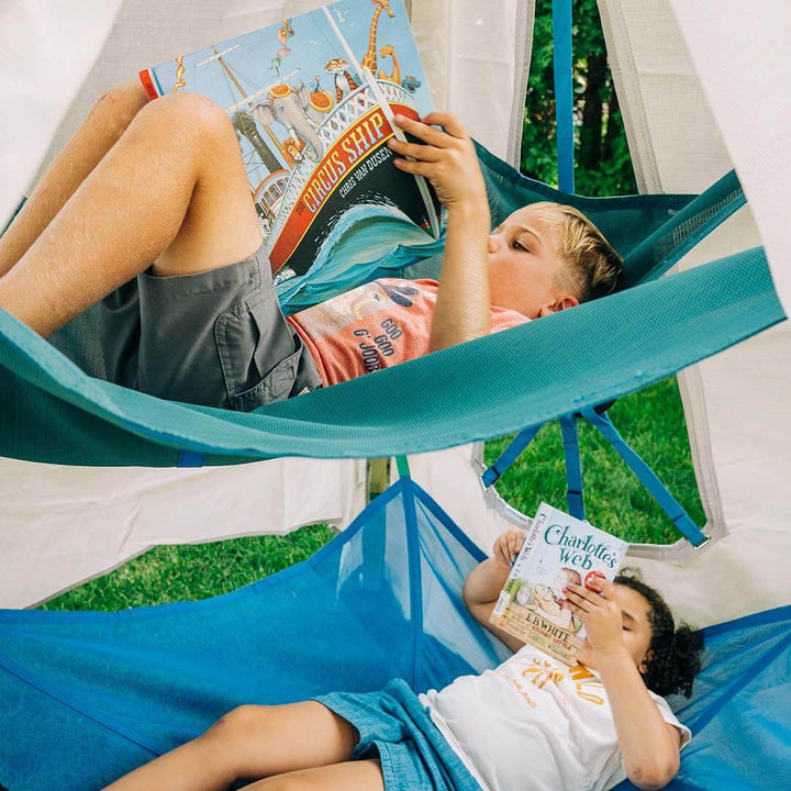 Two children relax in separate hammocks strung between a white canopy tent outdoors on grass. The child in the upper teal hammock holds a book while the child in the lower blue hammock.