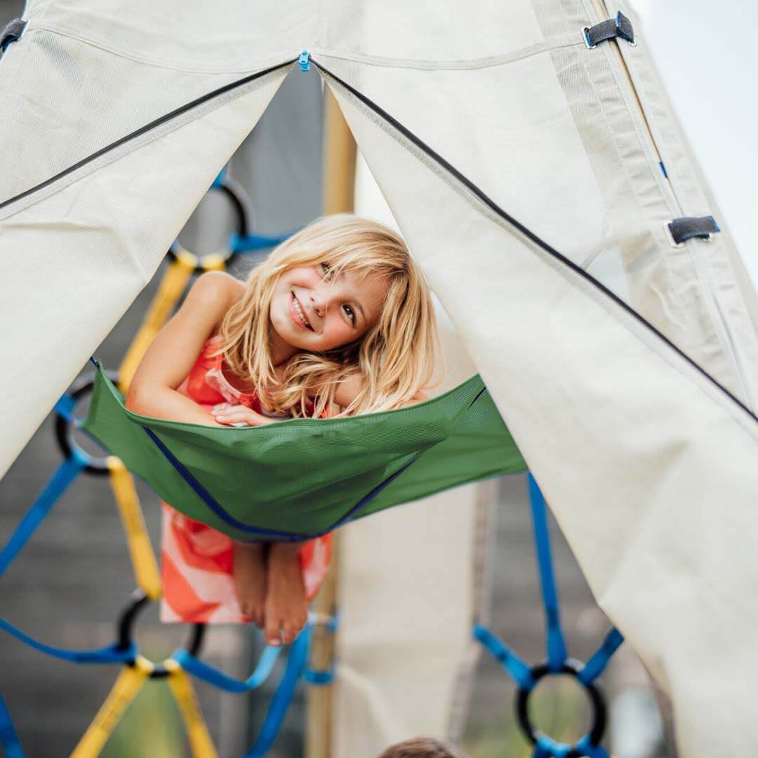 A young girl with blonde hair smiles while lying in a green fabric hammock suspended inside a beige play tent at an outdoor playground.