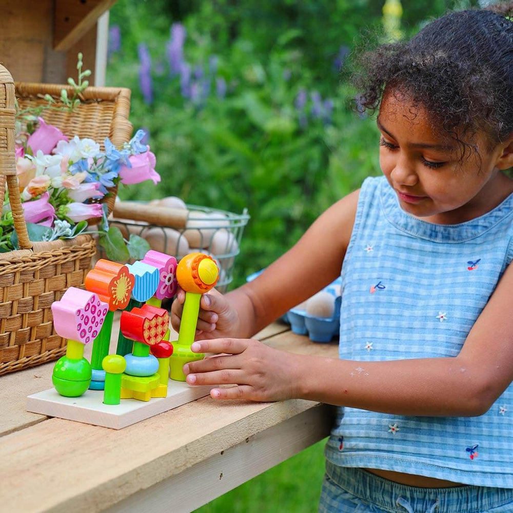 A young girl with curly hair wearing a blue checkered sleeveless top is playing with a colorful wooden toy set on a wooden table outdoors. The toy set features various bright-colored flower shapes.