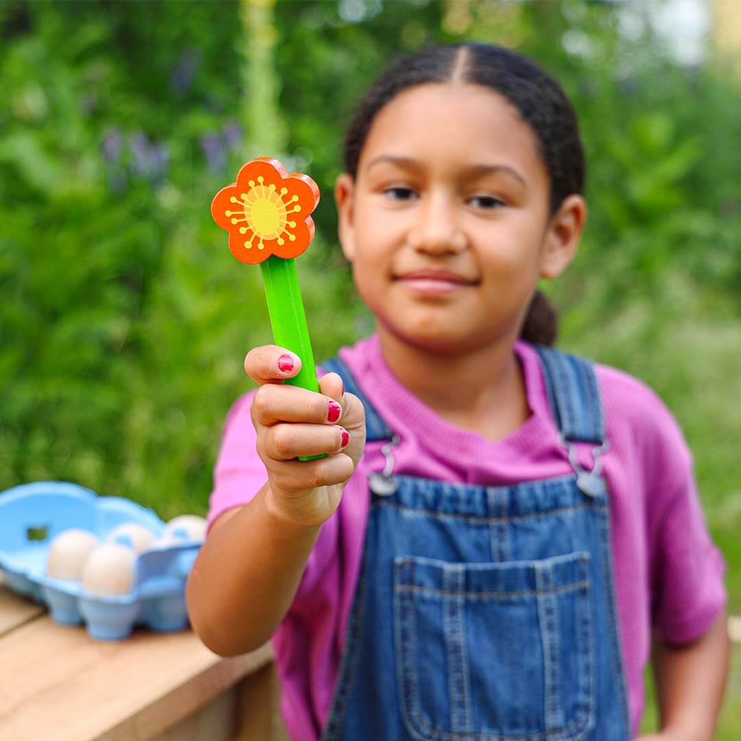 A young girl in a pink shirt and blue denim overalls holds up a small flower-shaped toy with an orange bloom, yellow center, and green stem, set against a blurred green garden background.