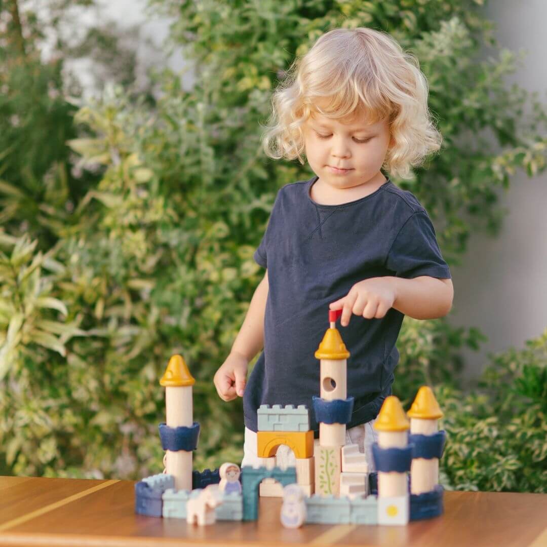 Child touching the red flag on the PlanToys wooden castle block set arranged as a fortress with blue and yellow towers, arches, walls, and small wooden figures including a king, queen, knight, and horse. Bella Luna Toys