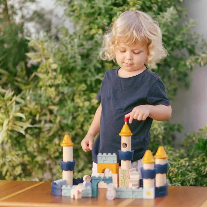 Child touching the red flag on the PlanToys wooden castle block set arranged as a fortress with blue and yellow towers, arches, walls, and small wooden figures including a king, queen, knight, and horse. Bella Luna Toys