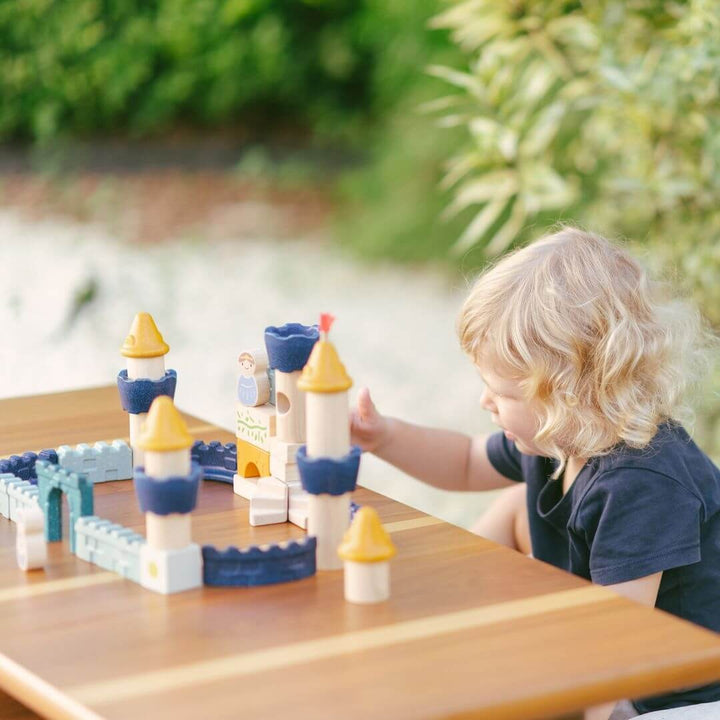 Child playing on a wooden table with the PlanToys wooden castle block set arranged as a fortress with blue and yellow towers, arches, walls, and small wooden figures including a king, queen, knight, and horse. Bella Luna Toys
