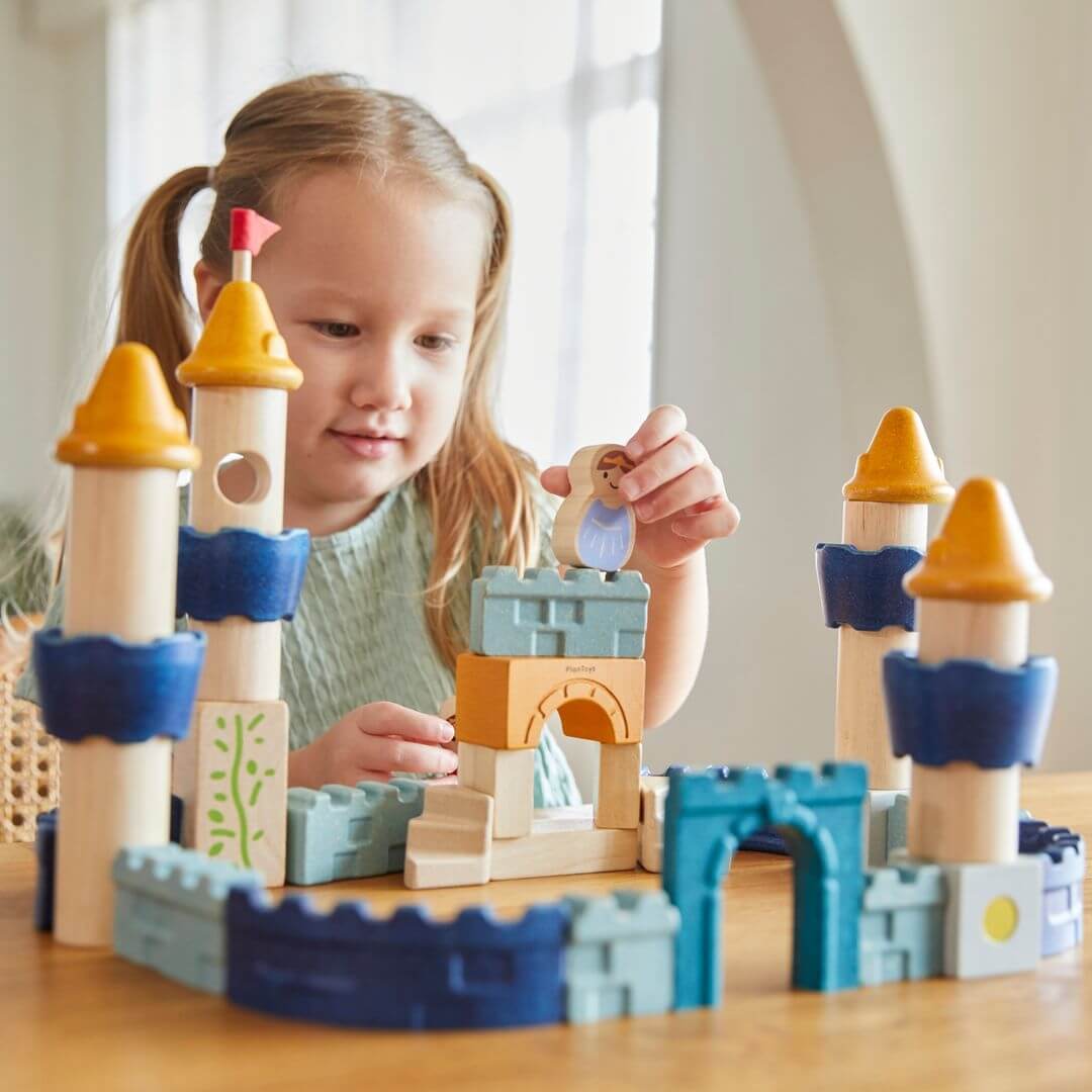 Child placing the queen wooden character on top of a turret in the PlanToys wooden castle block set arranged as a fortress with blue and yellow towers, arches, walls. Bella Luna Toys