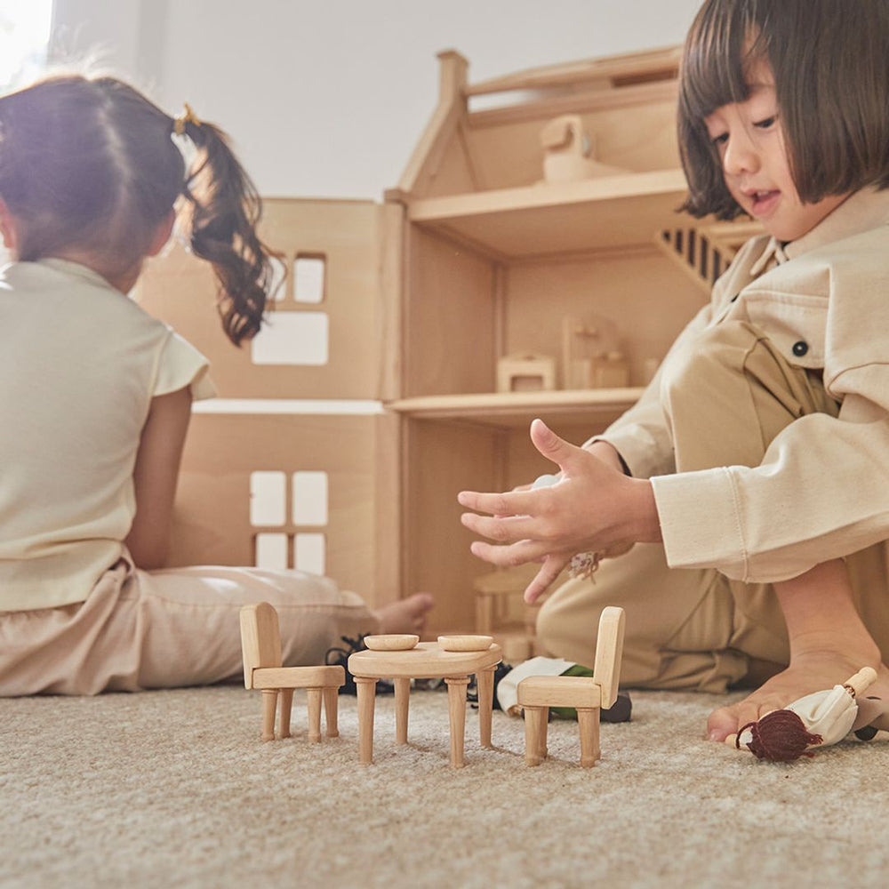 Two children playing with a fully furnished wooden dollhouse, arranging figures and furniture. The dining room table is set in front of one child on the beige carpet.